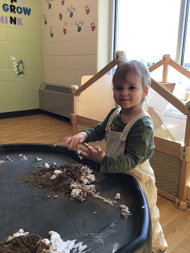 A child standing at a tuff tray playing with sand and shaving cream