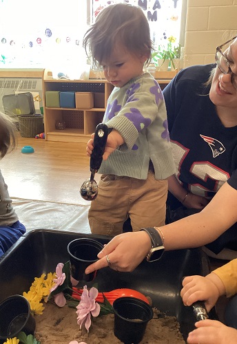 An infant standing in front of a sensory bin using a scoop to scoop up some sand