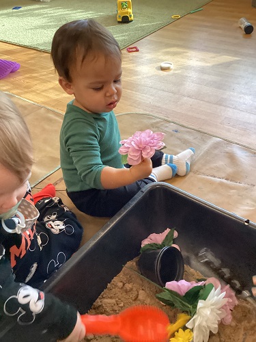 Two infants next to a sensory bin on the floor filled with sand, flowers, and scoops