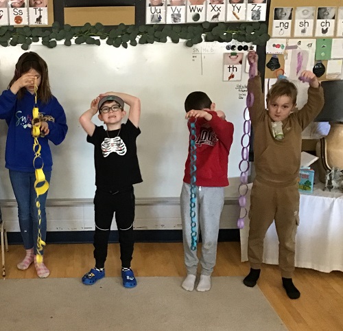 4 School Age children standing in front of the white board. 3 School Age children are holding a paper chain up, one School Age child has their arm in front of their face and the other School Age child is standing starring and smiling at the camera