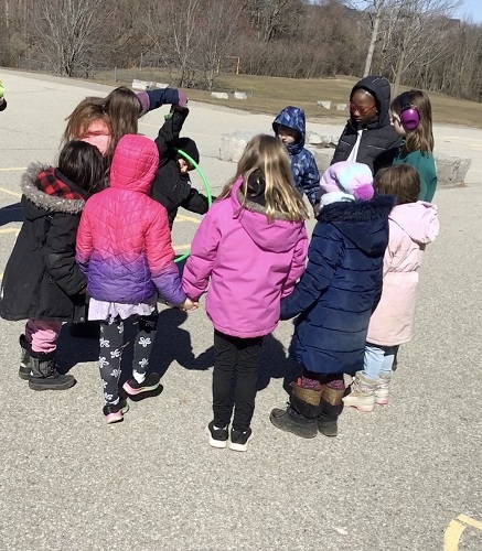 10 School Age children standing in a circle holding hands. One School Age child is holding another School Age child's arm up with a hula hoop in between them