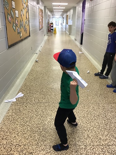 School Age child is standing against the wall looking at another School Age child who is standing in the middle of the hallway holding a paper airplane behind his head. You can see multiple paper airplanes sprawled out across the hallways floor