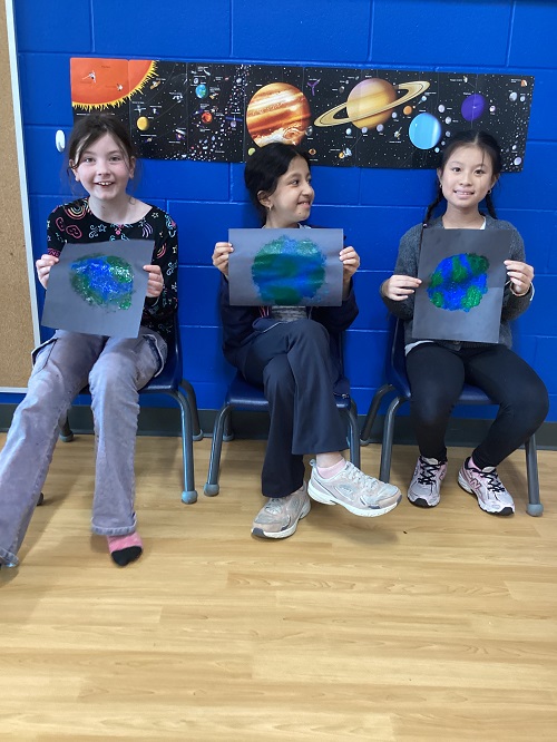 3 school age children siting on chairs holding up painting they did of earth