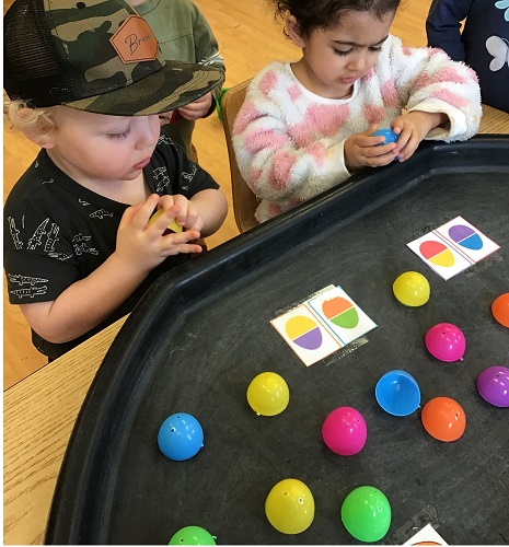 2 Preschool children are sitting at the tuff tray with various coloured eggs that are split apart with papers and different combination of coloured eggs. The 2 Preschool children are looking at the paper while holding an egg