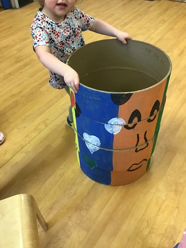 Preschool child standing holding a tube with painting on it that looks almost like a face, the child is looking at the camera and smiling