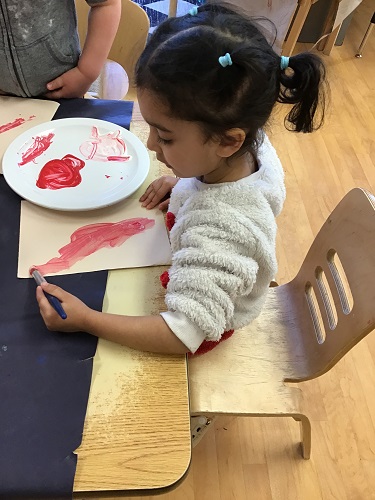 Preschool child sitting at a table with a plate of red paint, she is holding a paint brush and running it over the white paper in front of her. There is another Preschool child standing in the background.