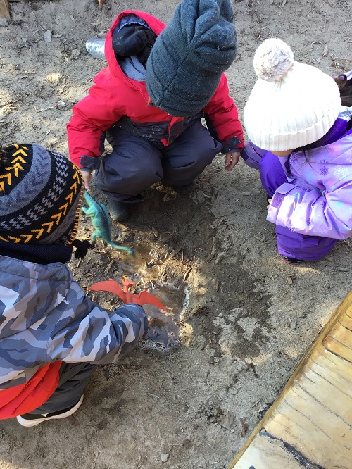 3 Toddler children outside crouching in a little circle and holding dinosaurs over top of a water puddle in the sand box