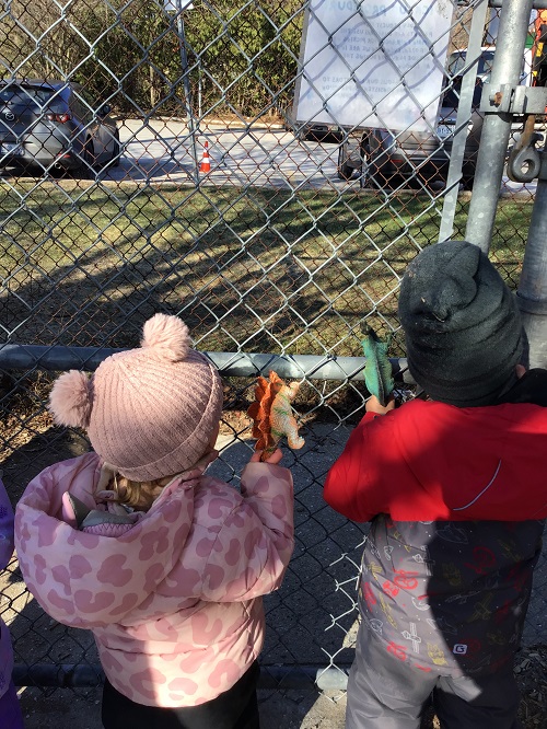 2 Toddler children standing at the gate holding onto a dinosaur in one hand and looking out at the cars in the parking lot