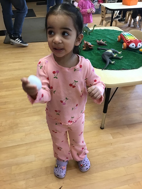 Toddler child standing in front of the camera looking off to the side holding a plastic white egg in her hand. There is another Toddler and an educator in the background just standing behind.