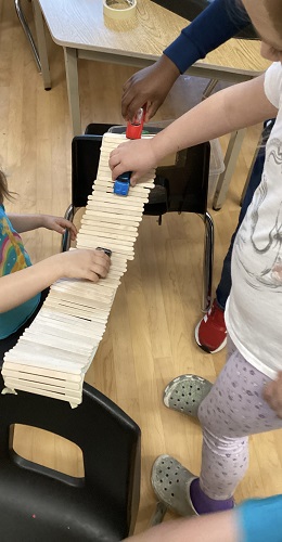 3 School Age children are standing, holding cars and driving them across a popsicle bridge that is being held up between 2 chairs