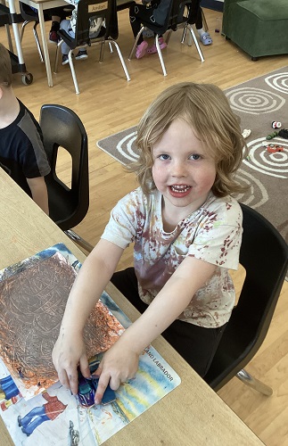 School Age child sitting at the desk holding one small car in each hand with a piece of paper with paint and car lines throughout, the child is looking up and smiling at the camer