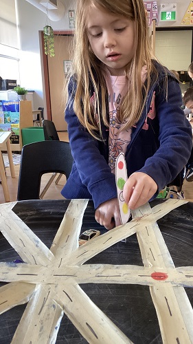 School Age child standing at the tuff tray with tape and broken lines, the child is holding an oval piece of paper that has yellow, ref, and green circles.