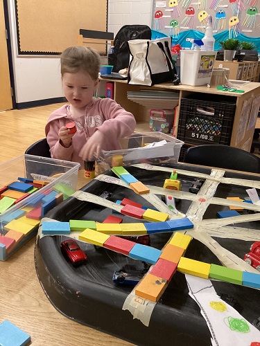 School Age child sitting at the table with a tuff tray in front of her with cars and taped roads. The child is holding a cylinder block in her hand and looking at it.