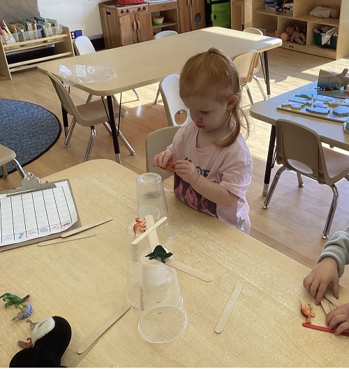 child playing with dinosaurs on a structure made out of plastic cups and popsicle sticks.